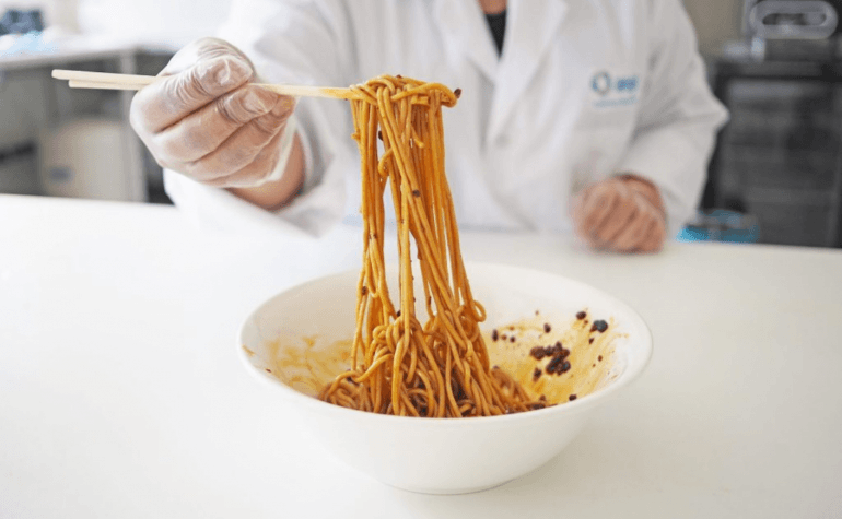 Gloved hand holding chopsticks over a bowl of oat noodles and sauce