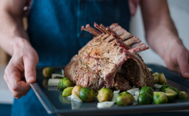 Hands holding baking dish with a cooked rack of lamb and vegetables